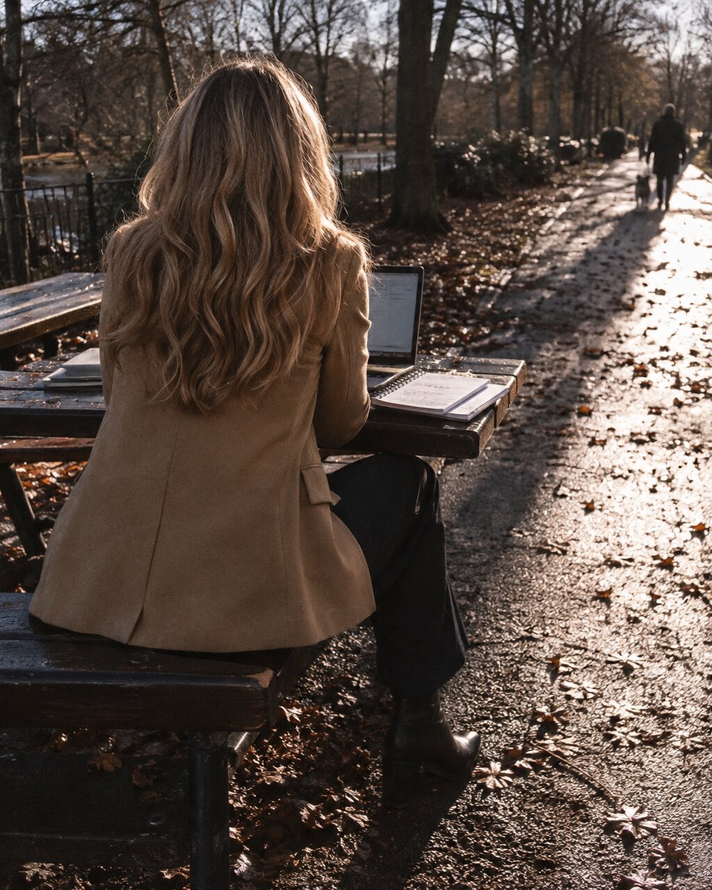 Woman seen from behind in a beige blazer using a laptop at a park bench