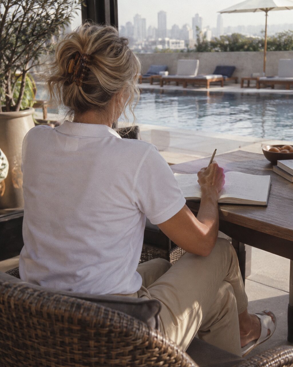 Woman seen from behind writing in a notebook beside a rooftop swimming pool