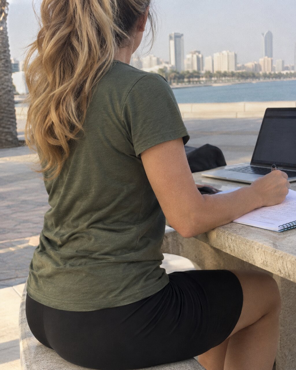 Woman seen from behind working on a laptop outdoors, overlooking a coastal city skyline