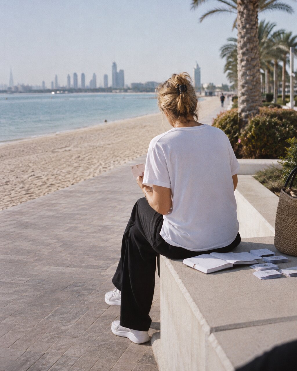 Woman seen from behind sitting on a wall by a beach, facing a distant skyline