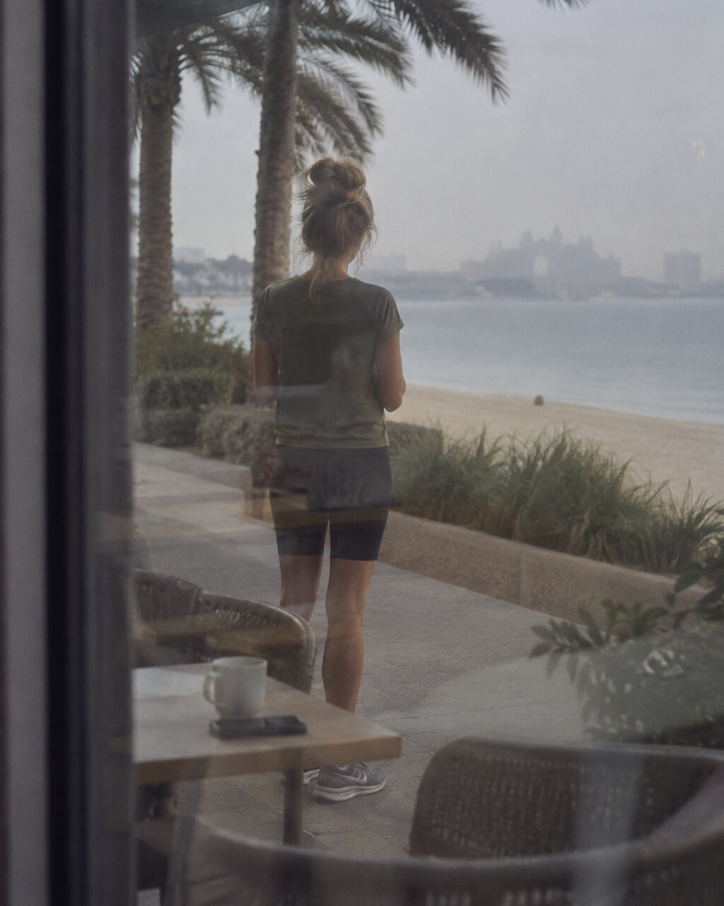 Woman seen from behind standing by a beach promenade, viewed through a cafe window
