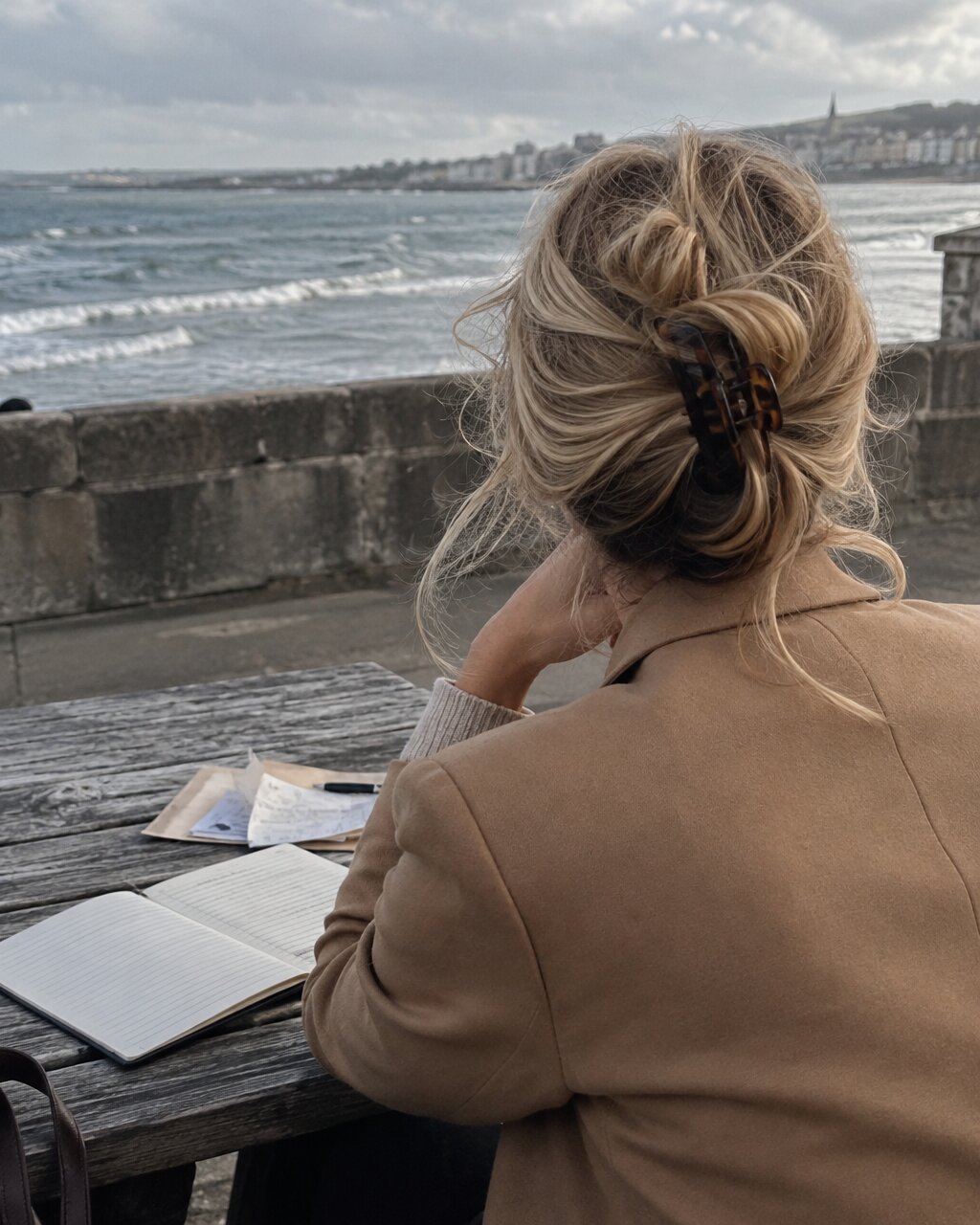 Woman seen from behind, sitting at a seaside wooden table with open notebooks