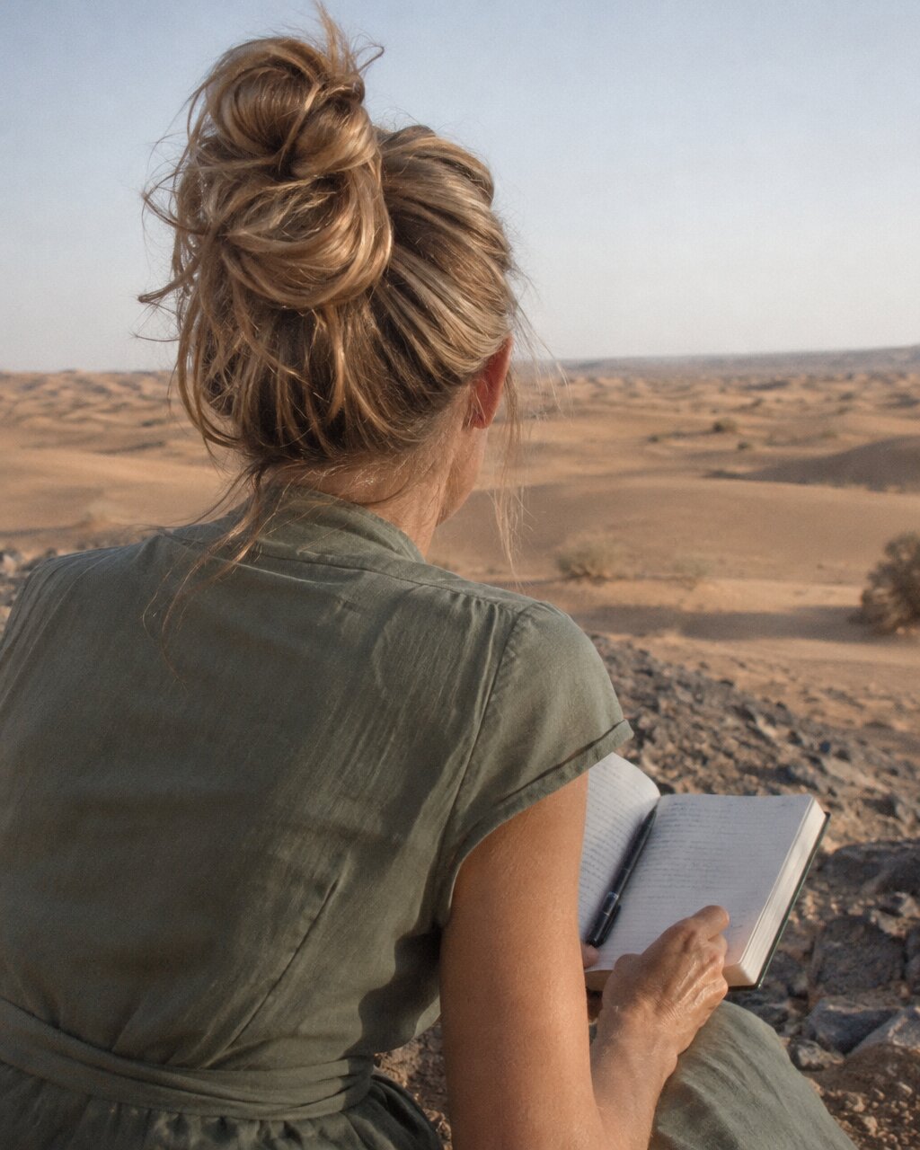 Woman seen from behind holding an open notebook and pen, overlooking a desert landscape