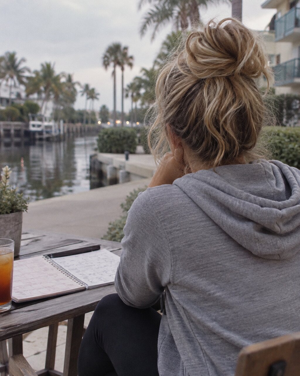 Woman seen from behind in a grey hoodie, sitting at a table overlooking a canal