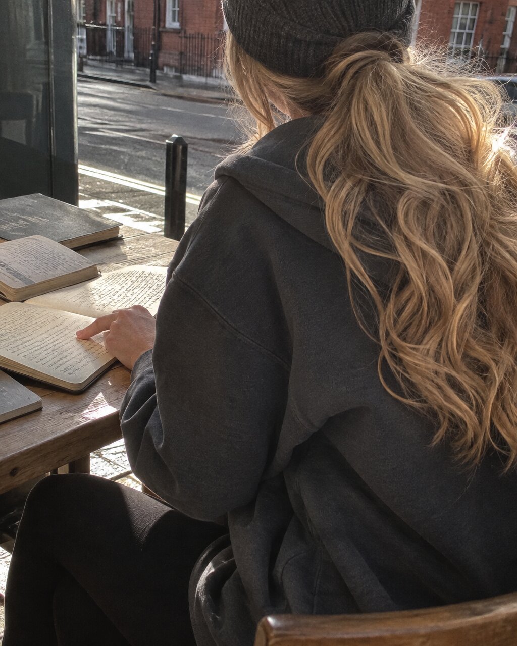 Woman seen from behind in a hoodie and beanie, reading at a window table